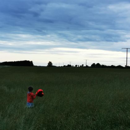 Een kind staat in een veld met hoge grassen en houdt een rood voorwerp vast. De lucht is bewolkt en er is voornamelijk vlakke landschap.
