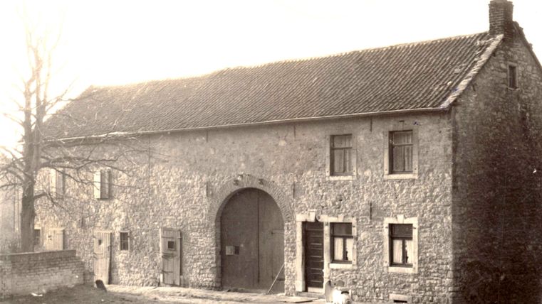 Een oud stenen huis met een grote poort en verschillende ramen. Op de voorgrond zijn enkele bomen en een vrijgemaakte weg te zien.