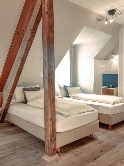 Een gezellige slaapkamer met twee eenpersoonsbedden en houten balken. Natuurlijke licht valt door het raam en er is een kleine televisie in de kamer.