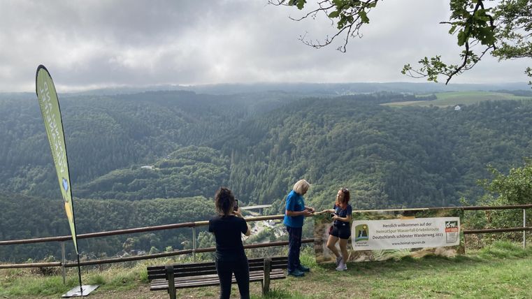 Eine Gruppe von Personen steht an einem Aussichtspunkt mit Blick auf eine hügelige Landschaft. Im Hintergrund sind bewaldete Hügel und ein bewölkter Himmel zu sehen.