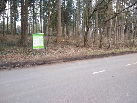 A signpost stands by the roadside in a forested area. The surroundings are quiet and green, with trees in the background.