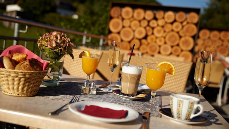 A laid table on a terrace with fresh pastries, juices, and coffee. In the background, garden furniture and wooden logs are visible.