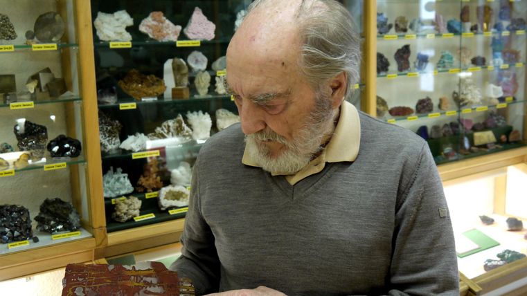 Ein älterer Mann betrachtet einen Mineralstein in einem Ausstellungsraum. Im Hintergrund sind viele weitere Mineralien und Steine zu sehen.