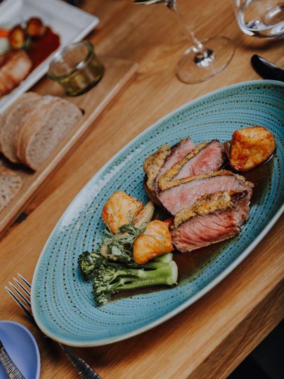 Ein kunstvoll angerichteter Teller mit zartem Fleisch, Kartoffeln und Brokkoli. Im Hintergrund sind Brot und ein Glas Wein zu sehen.