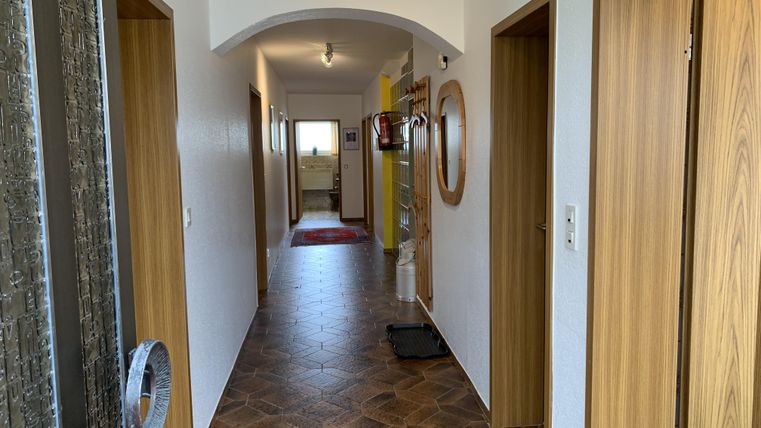 Ein Flur mit Holztüren und einem gemusterten Boden. Am Ende des Flurs ist ein heller Raum mit Fenster und einem Teppich.