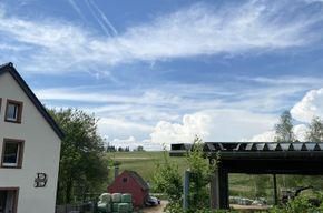 Ein hübsches ländliches Szenario mit einem weißen Haus und einer grünen Landschaft. Der Himmel ist blau mit weißen Wolken und einigen Flugzeugspuren.