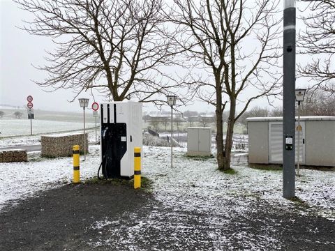 A charging station for electric cars is set in a snow-covered environment. In the background, trees and some signal and light poles can be seen.