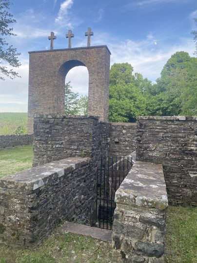Steinernes Kriegerdenkmal in Stadtkyll mit drei Kreuzen auf einem Torbogen, umgeben von grünen Bäumen und blauem Himmel.