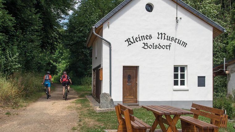 White building with the inscription 'Kleines Museum Bolsdorf'. Two cyclists on a path beside it. Wooden table and benches in the foreground.