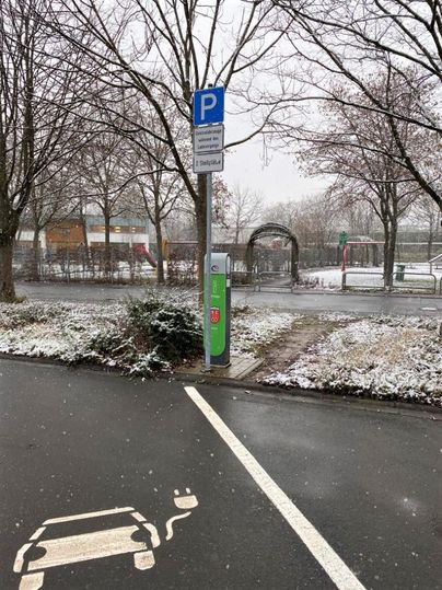A parking lot with a sign and a charging station for electric cars. There is snow, and the trees in the background are wintery bare.