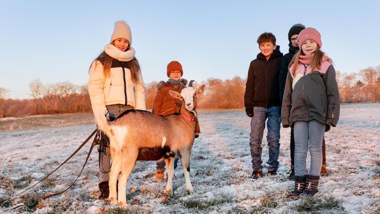Eine Gruppe von Kindern und Jugendlichen steht auf einem verschneiten Feld. Sie lächeln und sind mit einer Ziege beschäftigt.