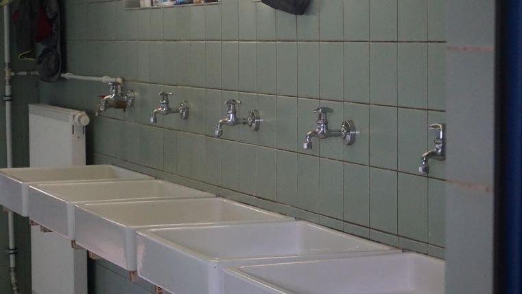 Ein Waschraum mit sechs Wasserhähnen und Waschbecken an einer gefliesten Wand. Oben sind Fenster und einige persönliche Gegenstände.