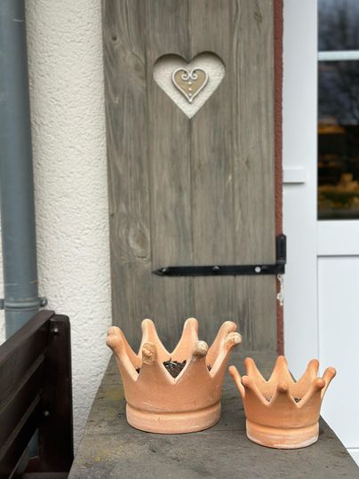 Twee aardewerken vaten in de vorm van kronen op een houten oppervlak. Op de achtergrond is een deur met een hartvormige uitsnede zichtbaar.