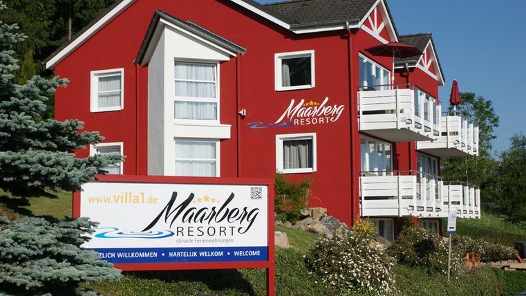 Een rood gebouw met de naam "Mahnberg Resort" en verschillende balkons. Een welkomstbord staat vooraan, omringd door groene landschappen.