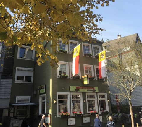 Ein grünes Gebäude mit großen Fenstern und zwei Fahnen. Im Vordergrund sind Passanten und herbstliche Blätter zu sehen.