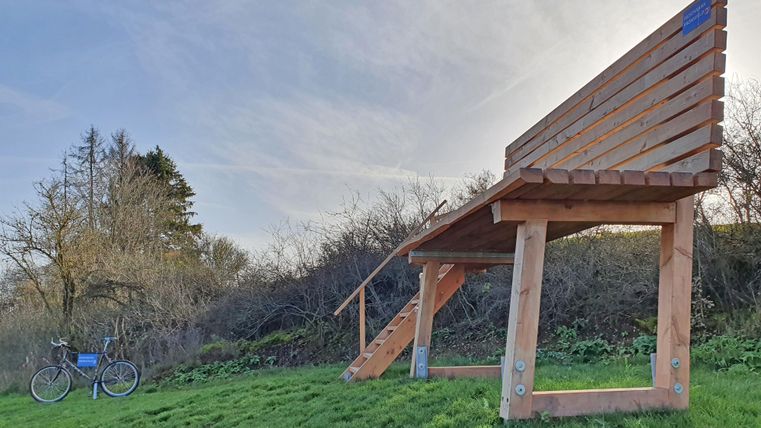 A large wooden bench stands on a meadow, next to it is a bicycle. In the background, there are trees and a clear sky.
