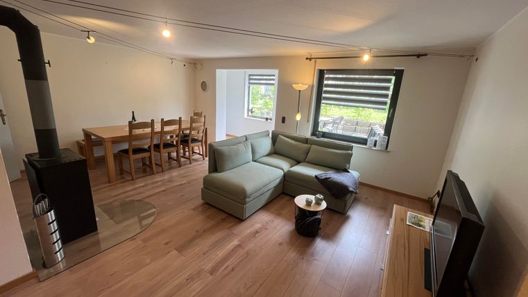A bright living room with a comfortable sofa and a fireplace. In the background, there is a dining table with chairs and large windows provide plenty of light.