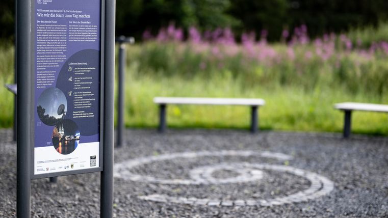 An information sign stands on a gravel area with a spiral marking. In the background, there are purple flowers and benches.