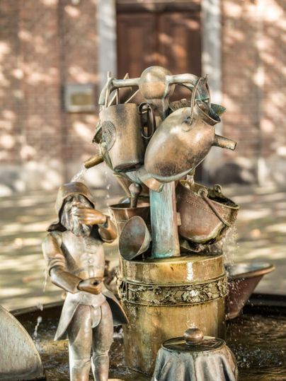 Eine kunstvolle Bronzeskulptur mit einem Soldaten und mehreren Kannen. Die Skulptur steht in einem Brunnen und reflektiert das Sonnenlicht.