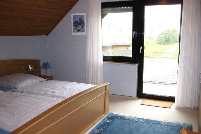 A cozy bedroom with a double bed and a wooden headboard. Large windows provide views of the terrace and a green outdoor area.