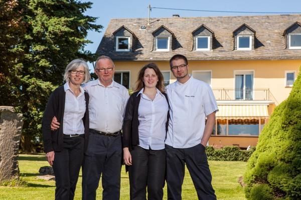 Een gezin van vier staat voor een groot, geel huis met een onderhouden tuin. Alle leden glimlachen en dragen lichte kleding.