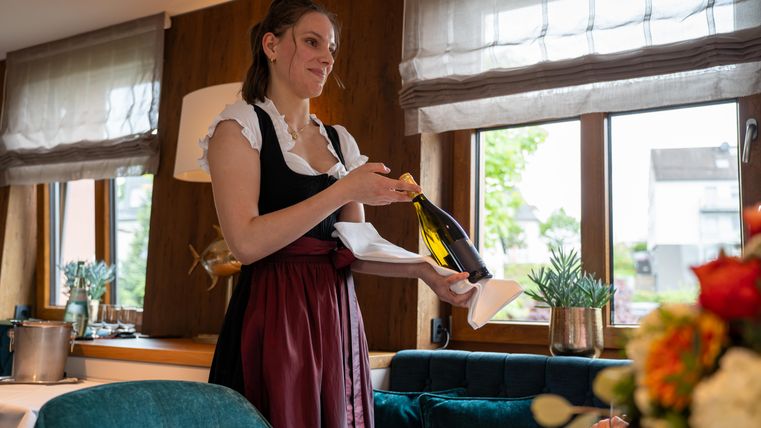 Een serveerster in traditionele klederdracht houdt een fles Champagne vast en serveert deze. Op de achtergrond zijn ramen met uitzicht naar buiten en een stijlvolle inrichting te zien.