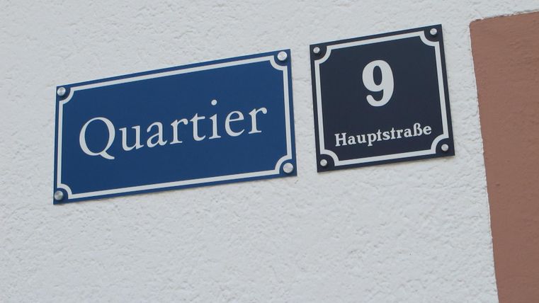 Das Bild zeigt ein Straßenschild mit der Aufschrift "Quartier" und der Nummer 9. Daneben steht "Hauptstraße".