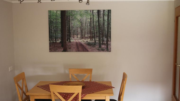 Een eetkamer met een houten tafel en stoelen. Aan de muur hangt een groot schilderij van een bosbedekte pad.