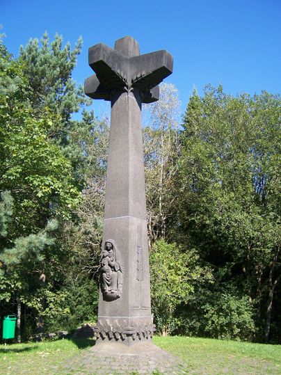 A tall monument in the shape of a cross stands in a green environment. The statue features a figurative representation at the base of the memorial stone.