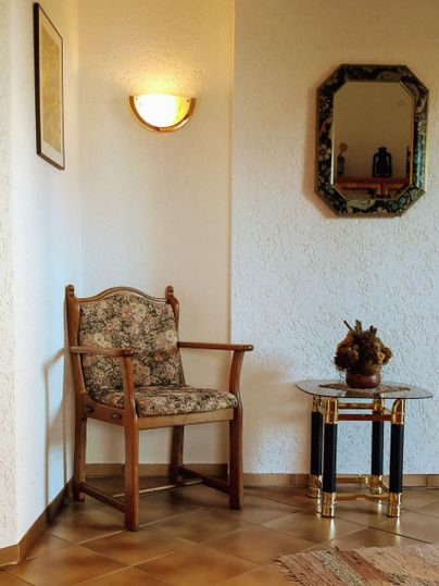 Een gezellige kamer met een stoel die is bekleed met een bloemenpatroon. Een kleine tafel met een vaas staat ernaast, en een spiegel hangt aan de muur.