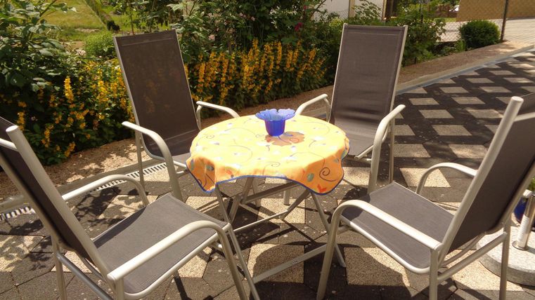 Ein runder Tisch mit einer bunten Tischdecke steht umgeben von vier Stühlen im Freien. Im Hintergrund sind Blumen und grünes Laub zu sehen.