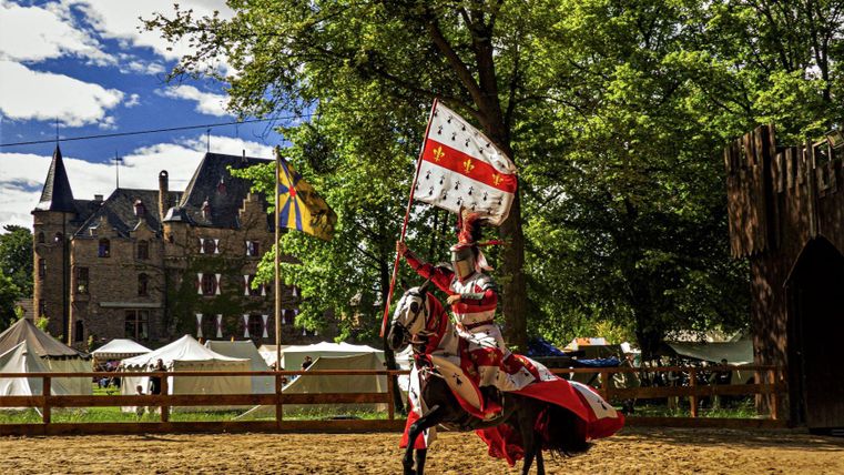 Een ridder in prachtige pantser rijdt op een paard en houdt een vlag vast. Op de achtergrond staat een historisch kasteel en middeleeuwse tenten.
