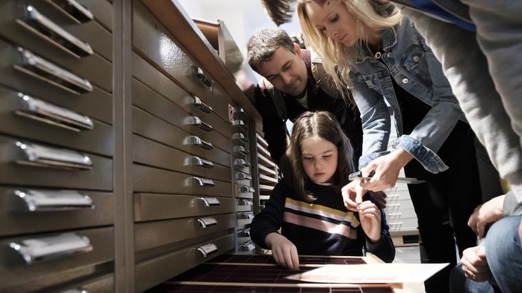 A group of people is attentively looking at a shelf with small compartments. A girl is exploring the objects inside, while the adults are helping her.