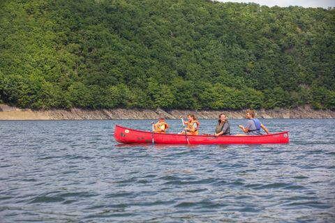 Een groep van vier personen peddelt in een rode kano op rustig water. Op de achtergrond is een groene, beboste kust te zien.