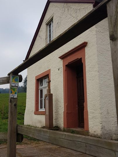 Ein kleines, weißes Haus mit roten Akzenten steht vor einer grünen Wiese. Die Fronttür ist offen und lädt ein, näher zu kommen.