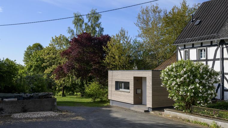 Ein moderner, holzverkleideter Anbau steht neben einem traditionellen Fachwerkhaus. Umgeben von Grün und Bäumen strahlt die Szene Ruhe und Gelassenheit aus.