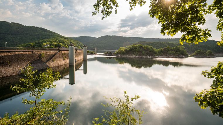 Ein ruhiger See mit Spiegelungen der umliegenden Berge und Wolken. Die sanften Wellen und das grüne Ufer verleihen der Szene eine friedliche Atmosphäre.