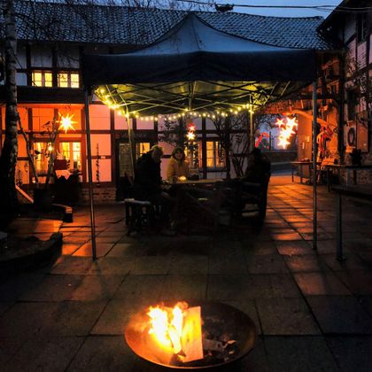 Een gezellige terras met een tentdak, waaronder mensen samenkomen. Op de voorgrond brandt een vuur in een metalen container, terwijl de omgeving verlicht is met warme lichten.