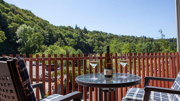 Ein gemütlicher Balkon mit einem Tisch und zwei Stühlen. Auf dem Tisch stehen eine Flasche Wein und zwei Weingläser, während die grüne Landschaft im Hintergrund zu sehen ist.