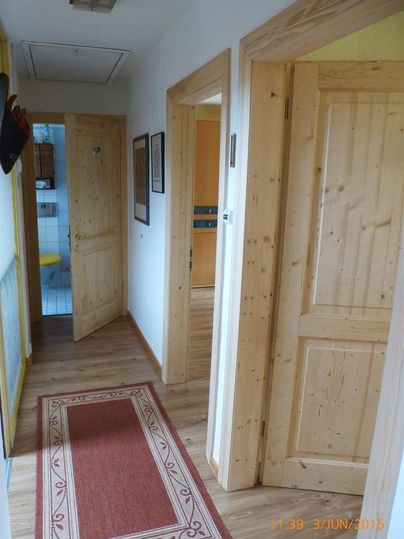 Ein heller Flur mit Holzvertäfelungen und braunem Laminatboden. An den Wänden hängen Bilder, und auf dem Boden liegt ein roter Teppich.