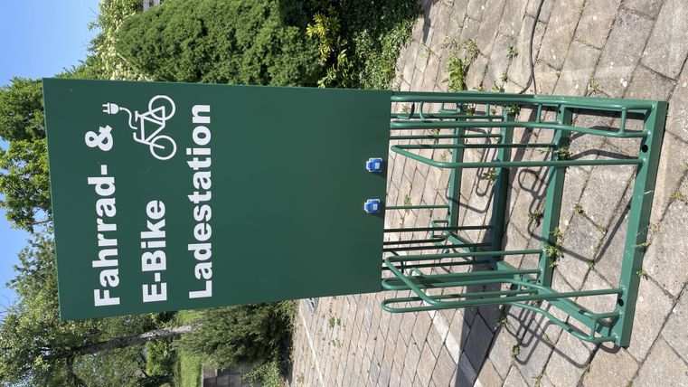 Eine Fahrrad- und E-Bike-Ladestation steht auf einem gepflasterten Bereich. Im Hintergrund sind Bäume und Sträucher sichtbar.