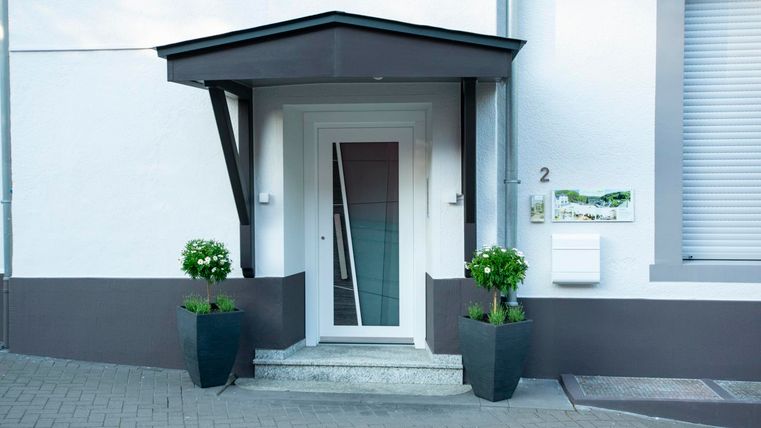 Ein moderner Hauseingang mit einer weißen Tür und einem überdachten Vordach. Zwei große Pflanzen in schwarzen Töpfen schmücken den Eingangsbereich.