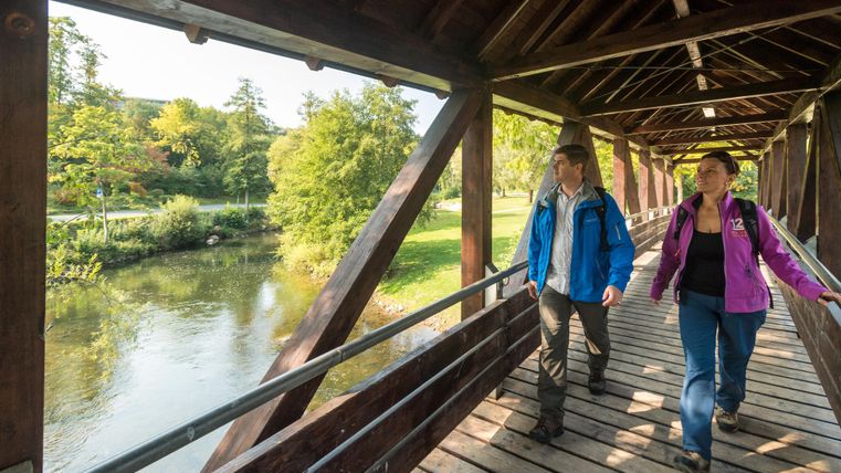 Zwei Personen gehen gemütlich über eine Holzbrücke. Im Hintergrund fließt ein ruhiger Fluss und die umgebende Natur zeigt herbstliche Farben.