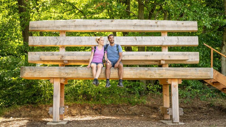 Ein großes Holzgestell im Freien, auf dem ein Paar sitzt. Es ist von grünen Bäumen umgeben und strahlt eine entspannte Atmosphäre aus.
