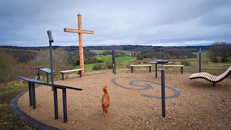 Een uitkijkpunt met een houten sculptuur in de vorm van een kruis. Op de voorgrond loopt een hond over het plein met uitzicht op de groene heuvels.
