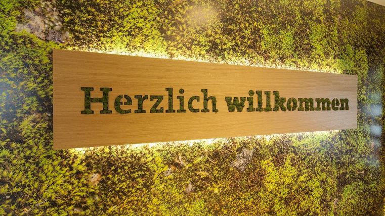 Eine Wand mit einem begrüßenden Schild aus Holz, das "Herzlich willkommen" sagt. Die Hintergrundfläche ist mit grünem Moos gestaltet.