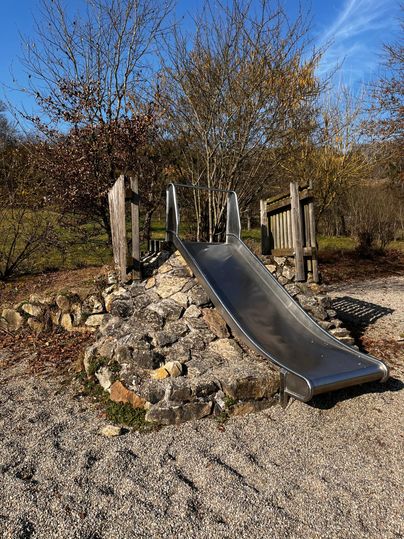 A slide made of stainless steel on a playground, surrounded by stones and gravel. In the background, trees and a blue sky are visible.