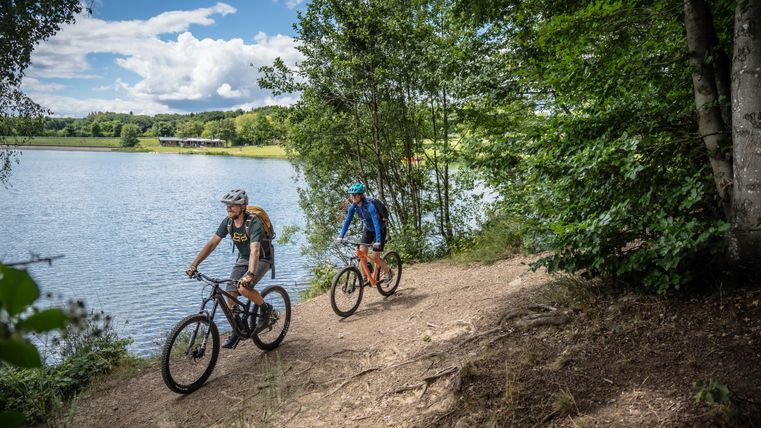 Zwei Radfahrer fahren am Ufer eines ruhigen Sees entlang. Umgeben von Bäumen und einer wunderschönen Landschaft.