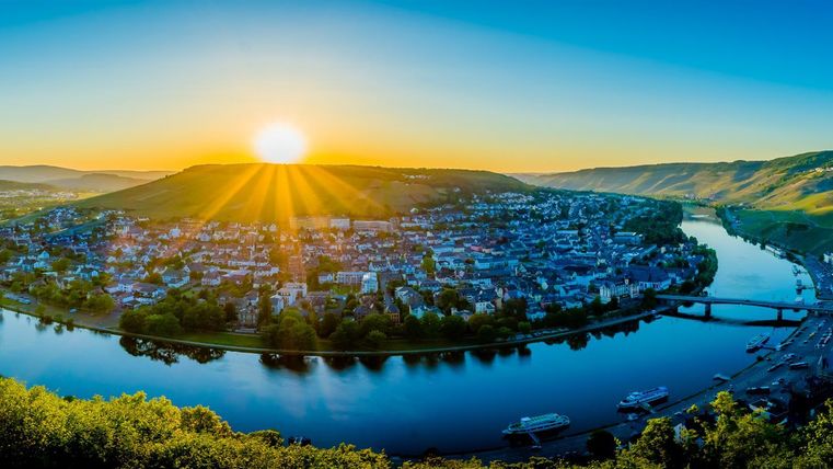 Eine malerische Stadtlandschaft am Fluss, umgeben von sanften Hügeln. Die Sonne geht hinter den Bergen auf und beleuchtet die Szene mit warmem Licht.