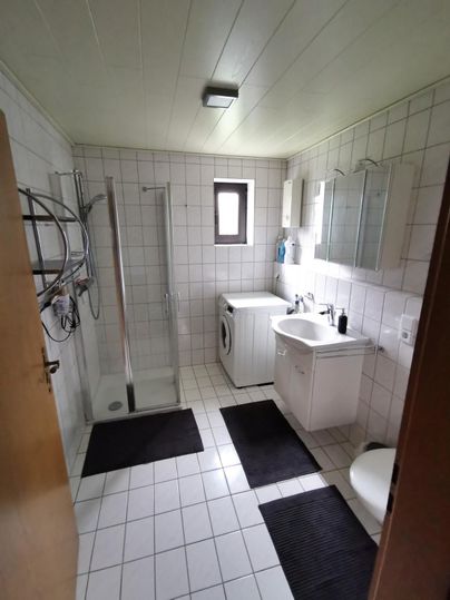 A modern bathroom with a shower, washing machine, and sink. The walls are tiled, and there are dark carpets on the floor.